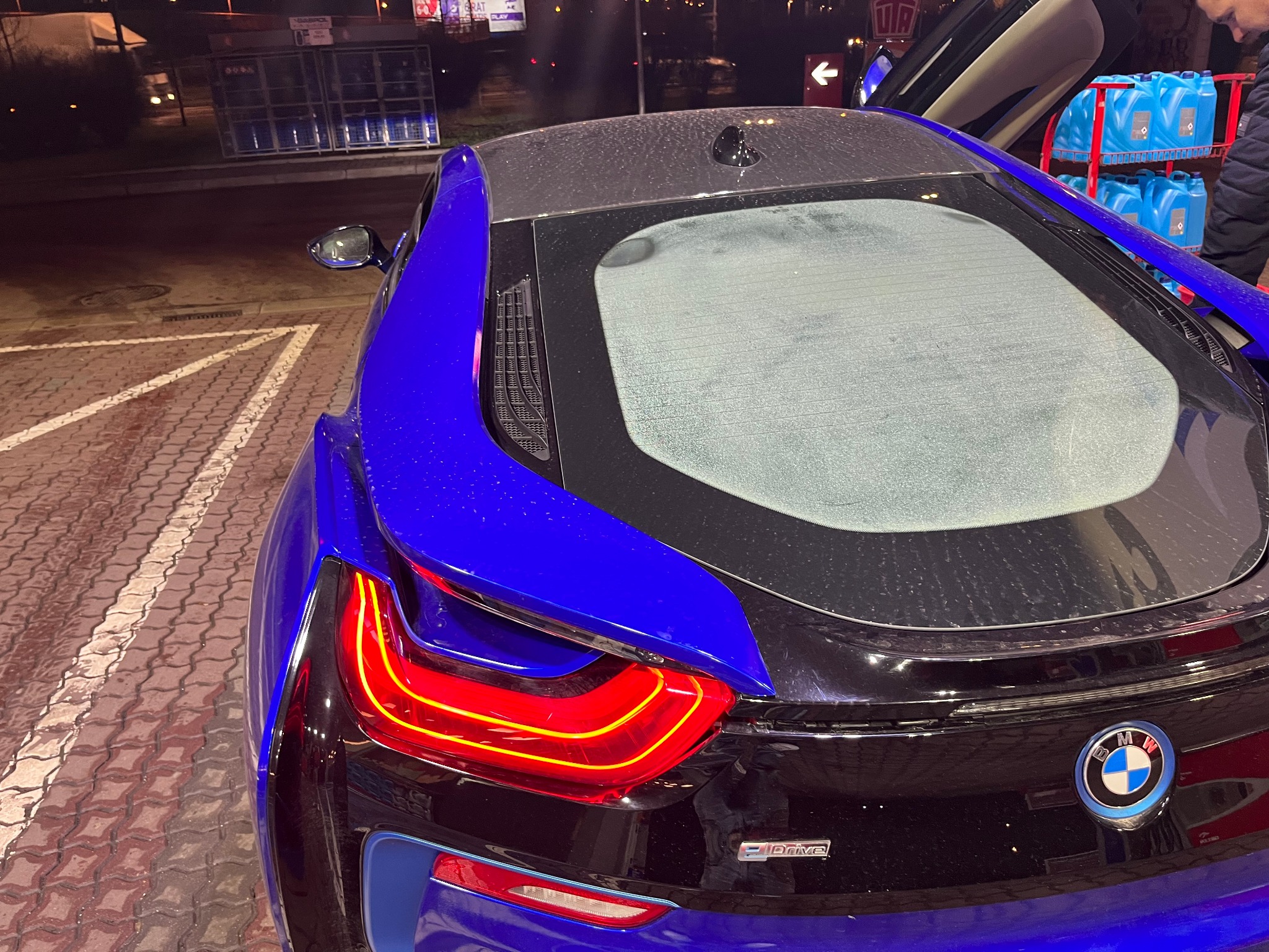 Tył niebieskiego BMW i8 z otwartymi drzwiami typu skrzydła, widoczne krople wody na karoserii i tylnej szybie, w tle kanistry z płynem do spryskiwaczy.