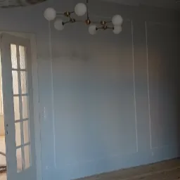 Wnętrze pokoju po malowaniu ścian na jasny kolor, widoczne świeżo pomalowane ściany z dekoracyjnymi listwami, drewniana podłoga, odkurzacz przemysłowy i białe, przeszklone drzwi.