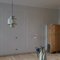 Jasne wnętrze z szarymi ścianami ozdobionymi białymi listwami, drewnianą podłogą, piecem kaflowym i nowoczesnym oświetleniem wiszącym oraz ściennym.