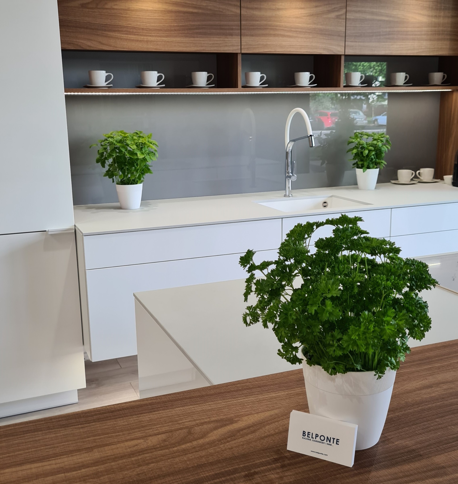 Nowoczesna kuchnia z białymi frontami i drewnianymi szafkami wiszącymi, akcentem zieleni w postaci ziół w doniczkach i wizytówką firmy Belponte na blacie.