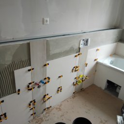 Remonty A-Z - Białe płytki układane na ścianie w łazience, widoczne krzyżyki dystansowe, ślady zaprawy, przygotowane miejsce na wannę, oraz puszka elektryczna.