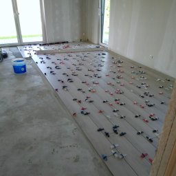 Remonty A-Z - Układanie podłogi z płytek imitujących drewno w jasnym pomieszczeniu, widoczne krzyżyki dystansowe i wiadro z klejem.