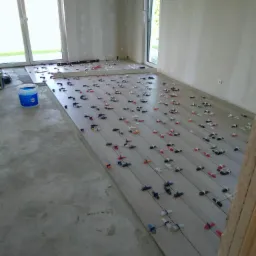 Układanie podłogi z płytek imitujących drewno w jasnym pomieszczeniu, widoczne krzyżyki dystansowe i wiadro z klejem.