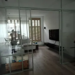 Przeszklone drzwi przesuwne z szarymi ramami oddzielające salon z drewnianą podłogą, jasną sofą i telewizorem na ścianie.