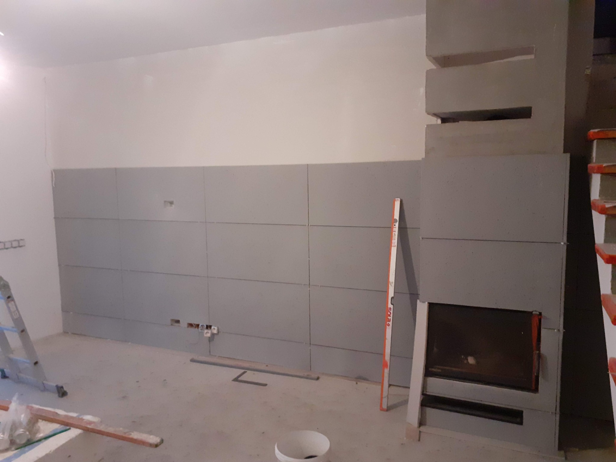 Wnętrze w trakcie montażu płyt karton gips na ścianie i obudowy kominka, widoczne profile, poziomica i drabina.