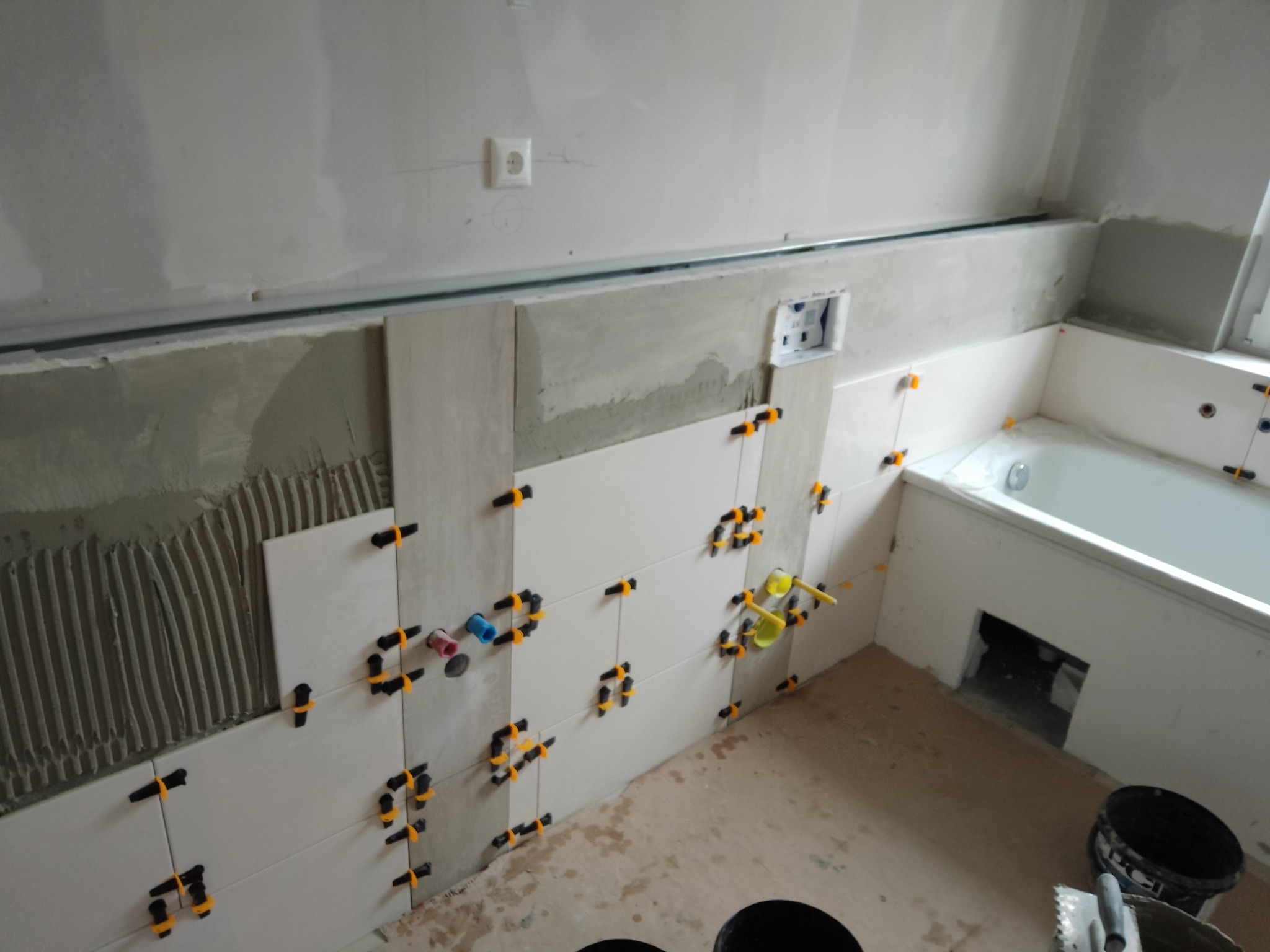 Białe płytki układane na ścianie w łazience, widoczne krzyżyki dystansowe, ślady zaprawy, przygotowane miejsce na wannę, oraz puszka elektryczna.