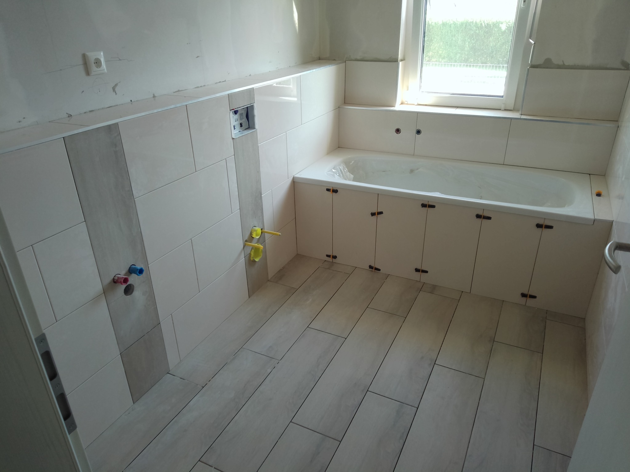 Wnętrze łazienki w trakcie wykańczania, widoczne ułożone płytki podłogowe imitujące drewno, częściowo wykończone ściany z jasnych płytek, zabudowana wanna z szafkami i wystające rury instalacji...