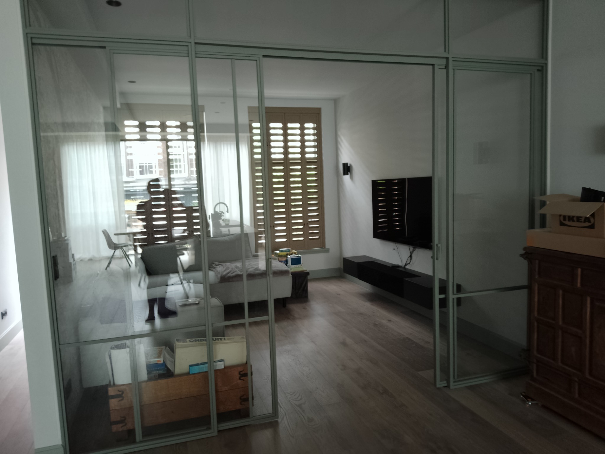 Przeszklone drzwi przesuwne z szarymi ramami oddzielające salon z drewnianą podłogą, jasną sofą i telewizorem na ścianie.