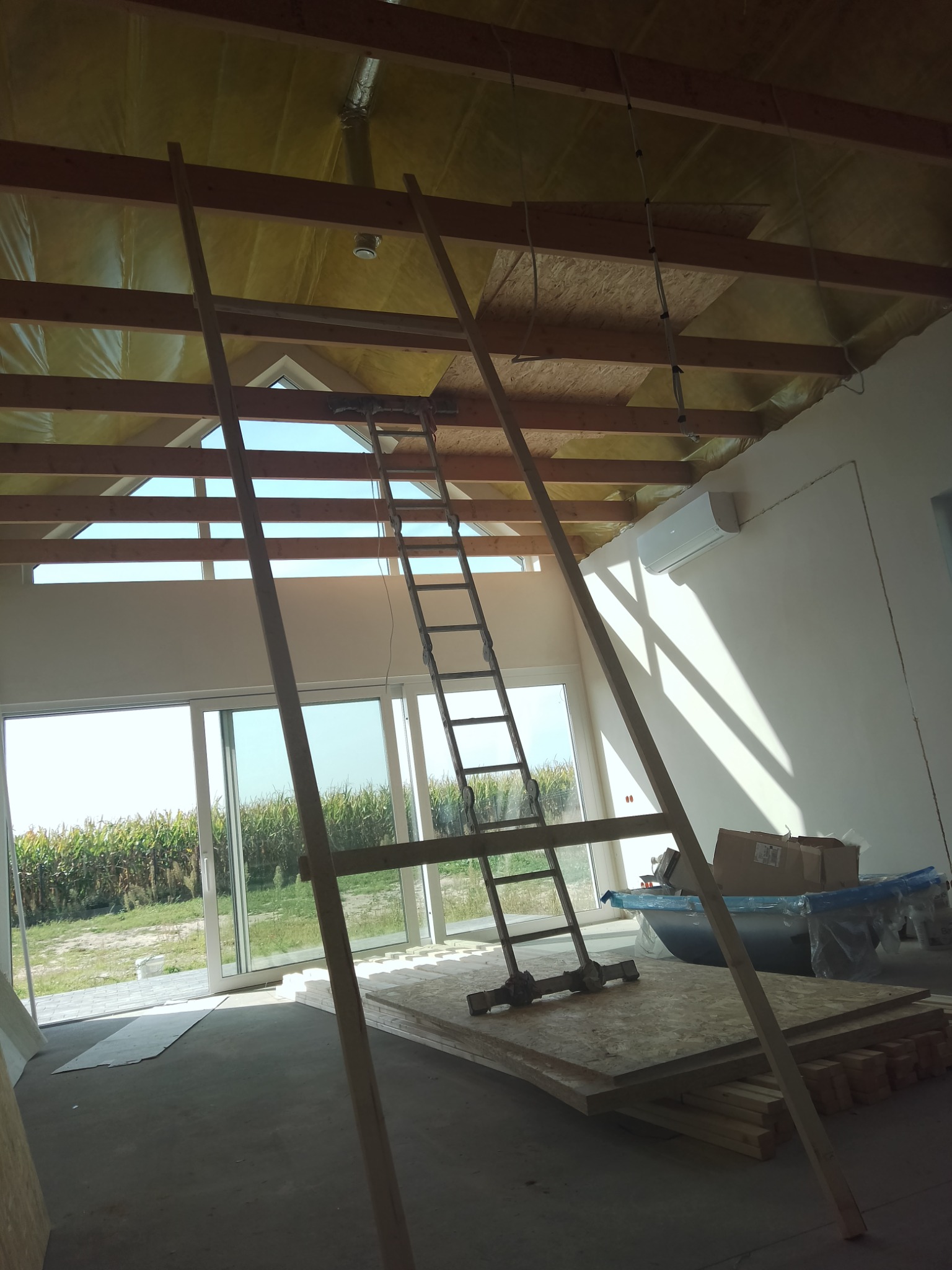 Wnętrze w trakcie remontu: widoczne ocieplenie dachu, drewniane elementy konstrukcyjne, drabina, płyty OSB, duże okno z widokiem na pole, klimatyzator na ścianie.