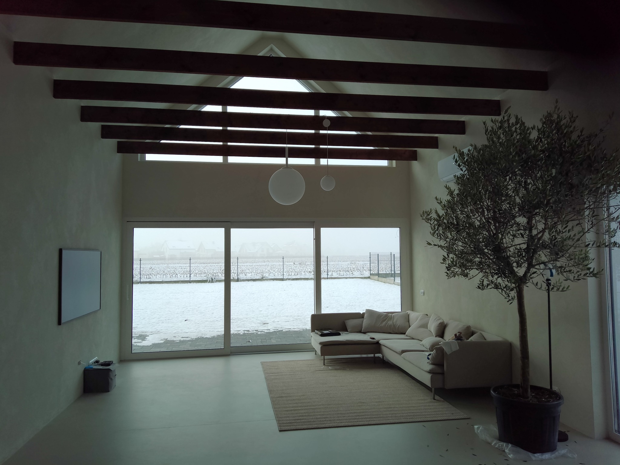 Jasne wnętrze z dekoracyjną gładzią na ścianach, drewnianymi belkami stropowymi, dużym oknem z widokiem na zaśnieżony krajobraz i drzewkiem oliwnym w donicy.