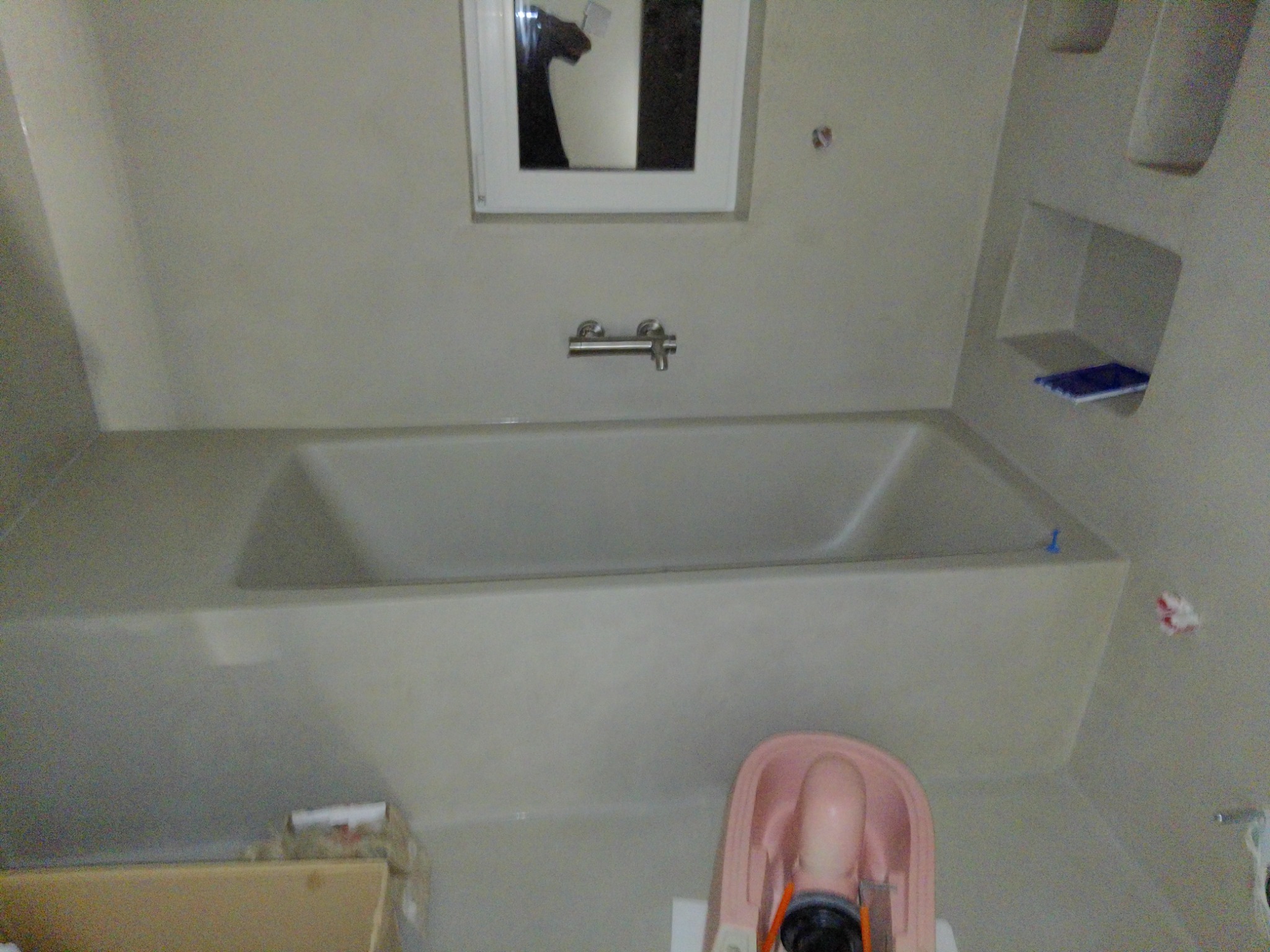 Nowoczesna, zabudowana wanna w łazience wykończonej w jasnych odcieniach szarości, z minimalistyczną armaturą i wbudowaną półką, widoczne akcesoria do montażu wanny.