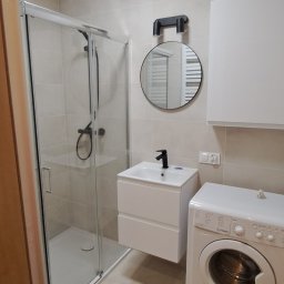 Oleksii Babak - Nowoczesna łazienka z kabiną prysznicową, umywalką z szafką, okrągłym lustrem z oświetleniem, pralką Indesit i beżowymi płytkami.