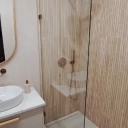 Nowoczesna łazienka z kabiną prysznicową typu walk-in, drewnopodobne panele ścienne, armatura w kolorze miedzi, umywalka nablatowa z szafką w odcieniach beżu i lustro w miedzianej ramie.