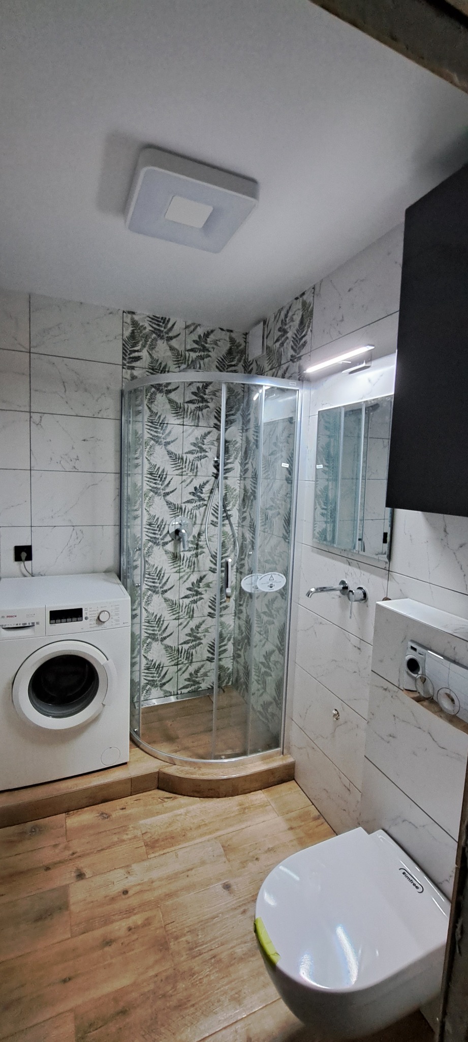 Ukończona łazienka z kabiną prysznicową narożną, pralką Bosch, podwieszaną toaletą i płytkami imitującymi marmur oraz z motywem paproci na ścianie prysznica.
