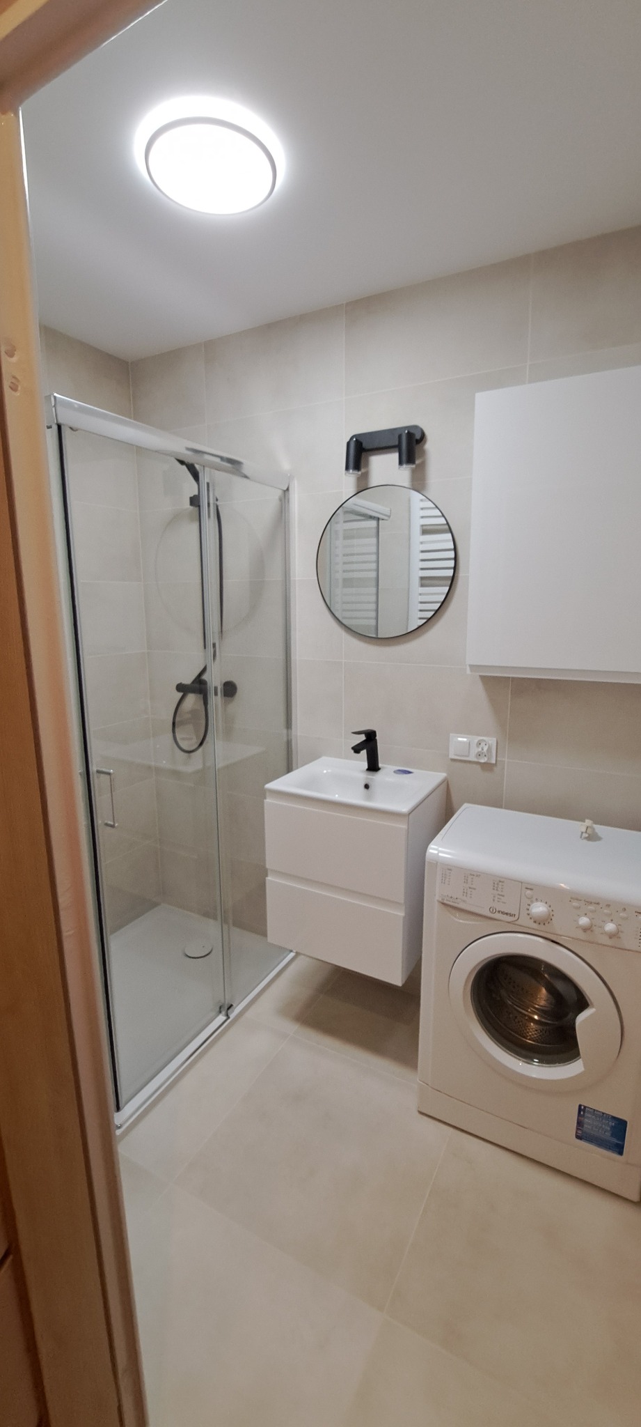 Nowoczesna łazienka z kabiną prysznicową, umywalką z szafką, okrągłym lustrem z oświetleniem, pralką Indesit i beżowymi płytkami.