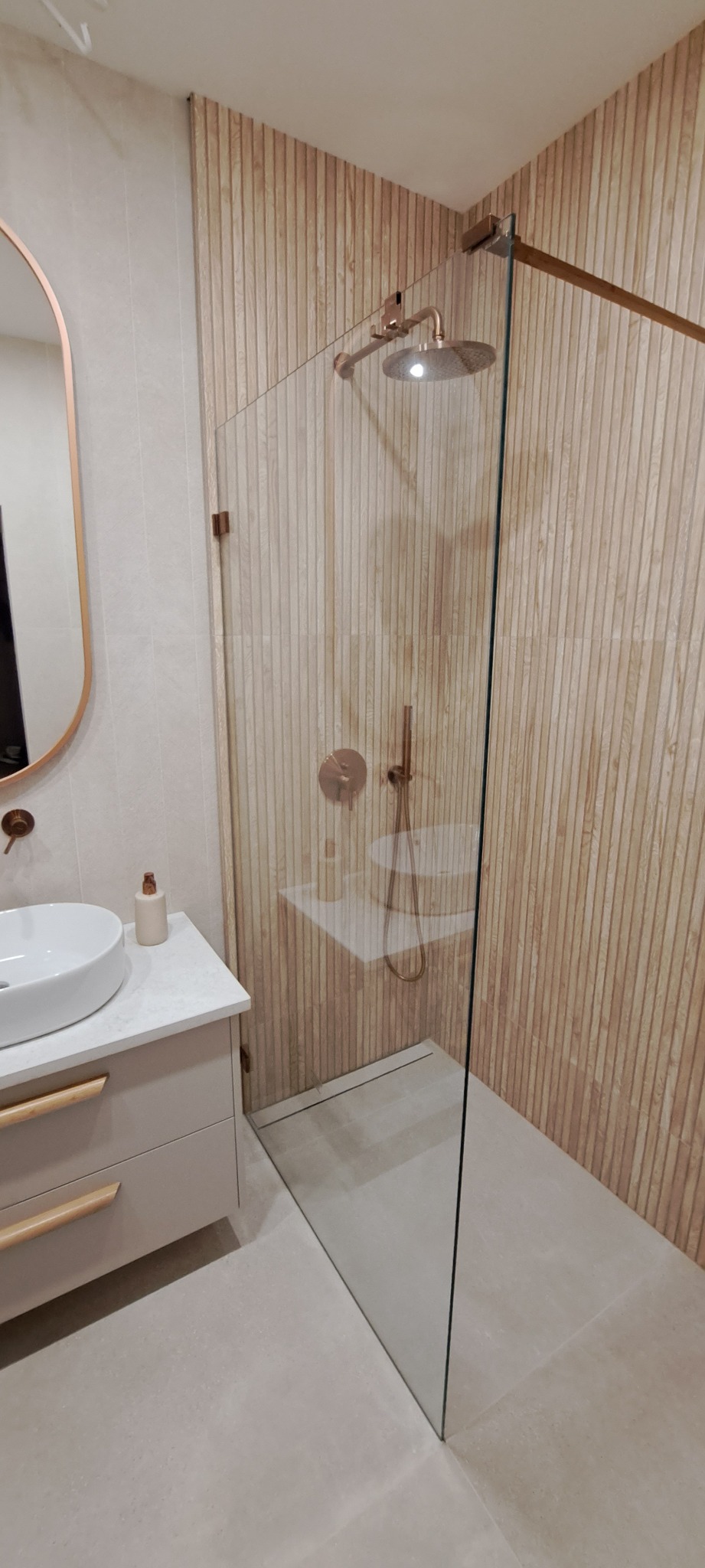 Nowoczesna łazienka z kabiną prysznicową typu walk-in, drewnopodobne panele ścienne, armatura w kolorze miedzi, umywalka nablatowa z szafką w odcieniach beżu i lustro w miedzianej ramie.