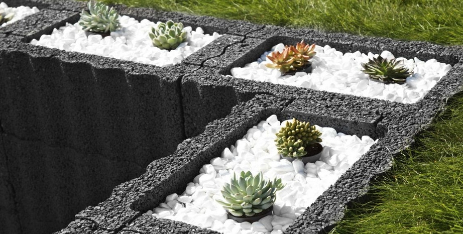 Kaskadowa rabata z szarych betonowych bloczków, wypełniona białymi kamieniami i różnymi sukulentami, widok z góry na tle trawy.