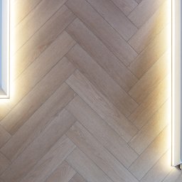 Bel-Pol partner Dekor. Podłogi Drzwi Panele Okna - Parkiet ułożony w jodełkę w odcieniach jasnego drewna, oświetlony liniowym światłem LED wzdłuż białej ściany, tworząc ciepły i nowoczesny efekt wizualny.