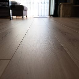 Bel-Pol partner Dekor. Podłogi Drzwi Panele Okna - Perspektywa z niskiego kąta na ułożoną, drewnianą podłogę z widocznym rysunkiem słojów i delikatnym połyskiem, w tle rozmyte elementy wyposażenia wnętrza i okno z zasłonami.