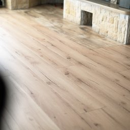 Bel-Pol partner Dekor. Podłogi Drzwi Panele Okna - Jasna, drewniana podłoga z widocznymi słojami i subtelnymi pęknięciami, ułożona wzdłużnie, z fragmentem kamiennej obudowy kominka w tle.