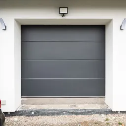 Szara, segmentowa brama garażowa z oświetleniem zewnętrznym i fragmentem zaparkowanego samochodu, widoczna na tle białej elewacji budynku.