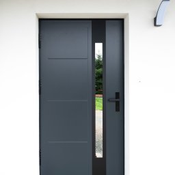 Bel-Pol partner Dekor. Podłogi Drzwi Panele Okna - Nowoczesne, grafitowe drzwi wejściowe z pionowym przeszkleniem, zamontowane w Kłodzku, z widocznym fragmentem ogrodu przez szybę i minimalistyczną lampą ścienną.