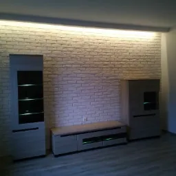 Ściana z białej cegły dekoracyjnej z podświetleniem LED, szafki i komoda w szarym odcieniu drewna, podłoga z paneli w odcieniach szarości.