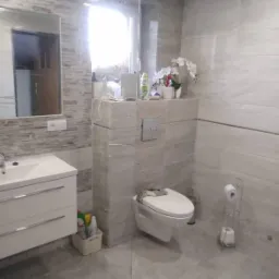 Nowoczesna łazienka wykończona w odcieniach szarości, z wiszącą toaletą z deską sedesową i podwieszaną umywalką z szafką, lustro z dekoracyjną ścianą imitującą kamień, akcesoria łazienkowe i kwiaty...