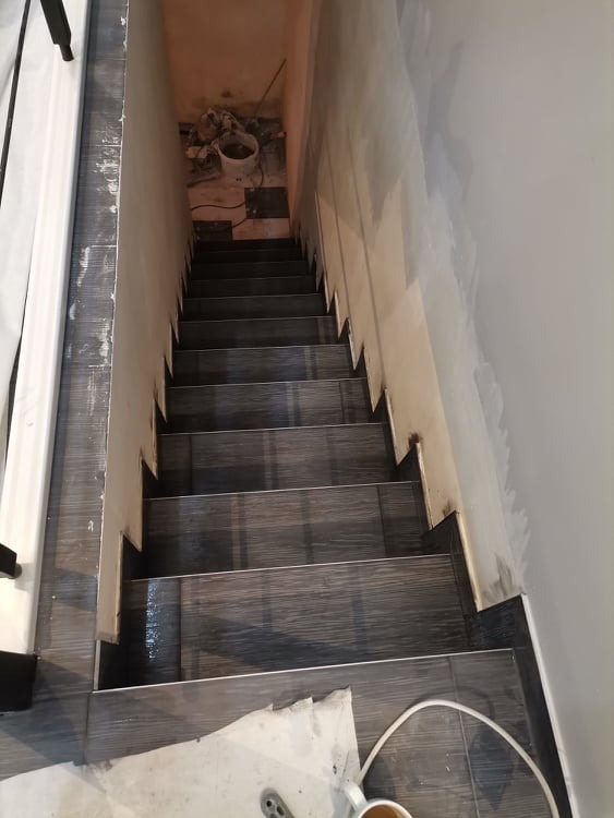 Schody wyłożone płytkami imitującymi drewno, widok z góry w trakcie prac wykończeniowych, ściany częściowo pomalowane, widoczne narzędzia i wiadro z zaprawą na górnym podeście.