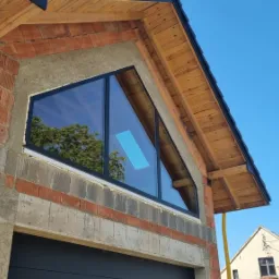 Trójkątne okno z czarną ramą w surowym stanie budynku, widoczne cegły i drewniana konstrukcja dachu, odbicie drzewa w szybie.