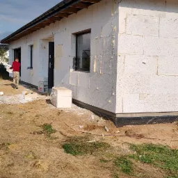 Dom jednorodzinny w trakcie ocieplania ścian zewnętrznych, widoczne białe bloczki i częściowo przyklejony styropian, pracownik przenosi materiały izolacyjne.