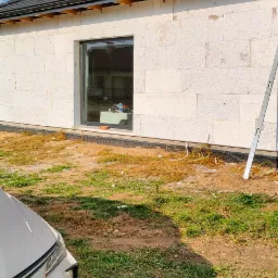 Surowa elewacja budynku z białych bloczków, widoczne okno z parapetem i akcesoria, częściowo widoczny samochód na pierwszym planie, trawa i ziemia wokół budynku.