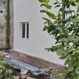 Fragment ściany budynku po tynkowaniu i malowaniu na biało, z widocznym oknem, rynną, elementami rusztowania i fragmentem starej, nieotynkowanej ściany obok.