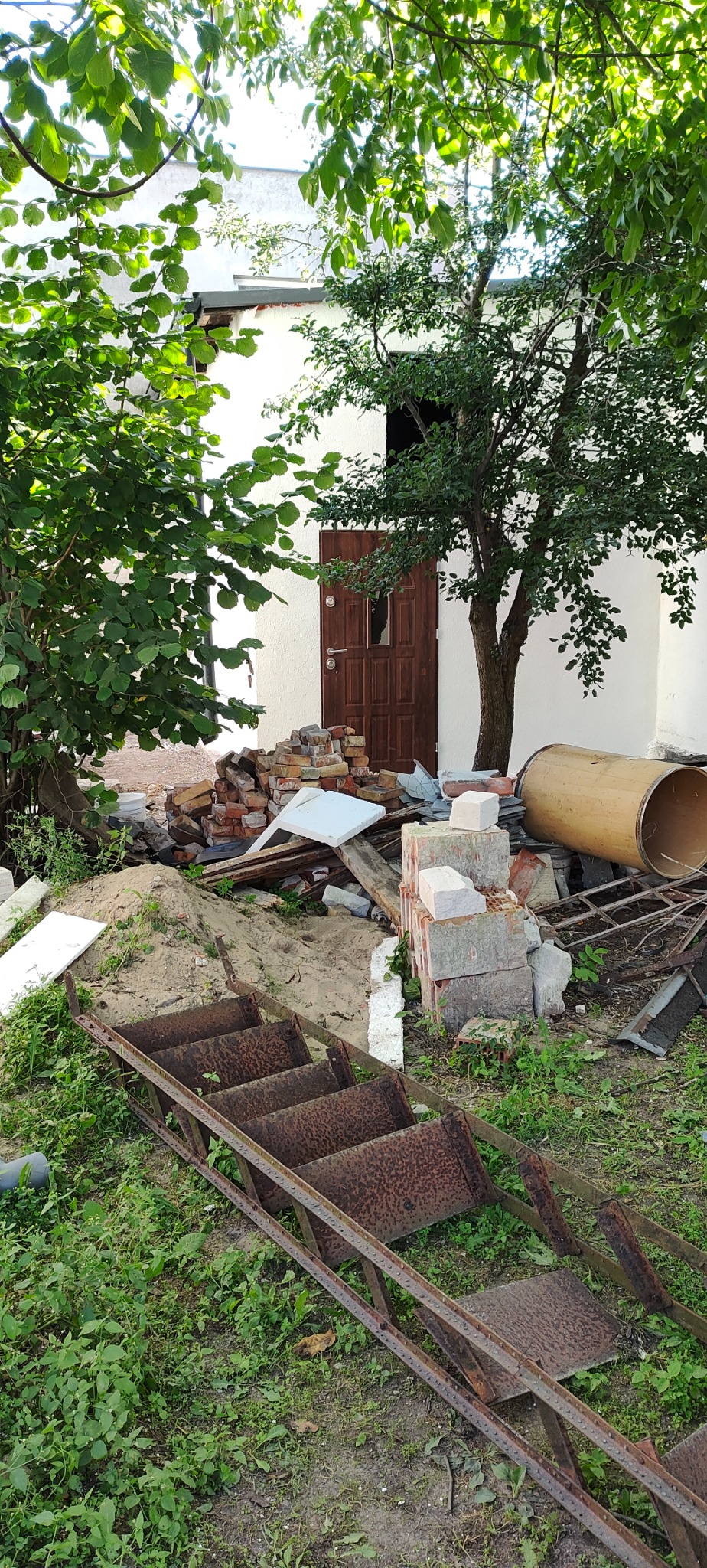 Przygotowania do prac murarskich w Wąbrzeźnie: sterta cegieł, piasku, pustaków i stara metalowa drabina oparta o białą ścianę budynku z drewnianymi drzwiami.