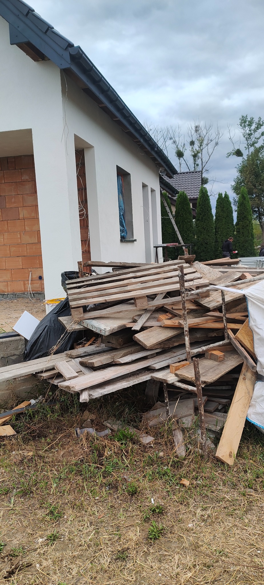 Dom w trakcie budowy: fragment otynkowanej i pomalowanej na biało elewacji, fragment ściany z czerwonej cegły, sterta drewna i gruzu na pierwszym planie, drabina oparta o ścianę, pochmurne niebo.