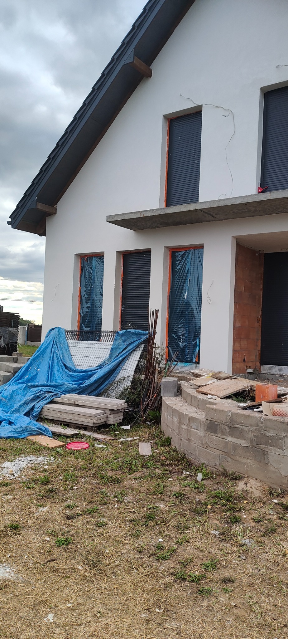Nowy dom w trakcie budowy, okna zabezpieczone folią, widoczne niedoskonałości tynku i elementy murowane, teren wokół zagracony materiałami budowlanymi i przykryty plandeką.