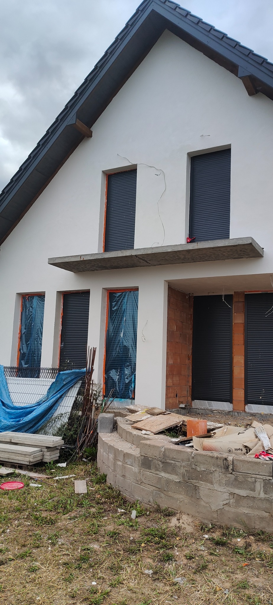 Dom jednorodzinny w trakcie prac elewacyjnych: widoczne ściany z nałożonym tynkiem, okna zabezpieczone folią, cegły i worki z zaprawą na pierwszym planie, wskazujące na trwające prace budowlane.