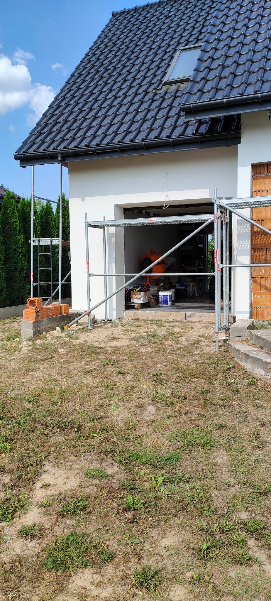 Dom jednorodzinny z białymi ścianami w trakcie malowania elewacji, widoczne rusztowania, cegły i sprzęt budowlany w otwartym garażu.