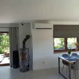 Nowoczesny salon z klimatyzatorem ściennym Gree, piecem wolnostojącym z rurą dymową i minimalistycznym stołem jadalnym przy oknie.