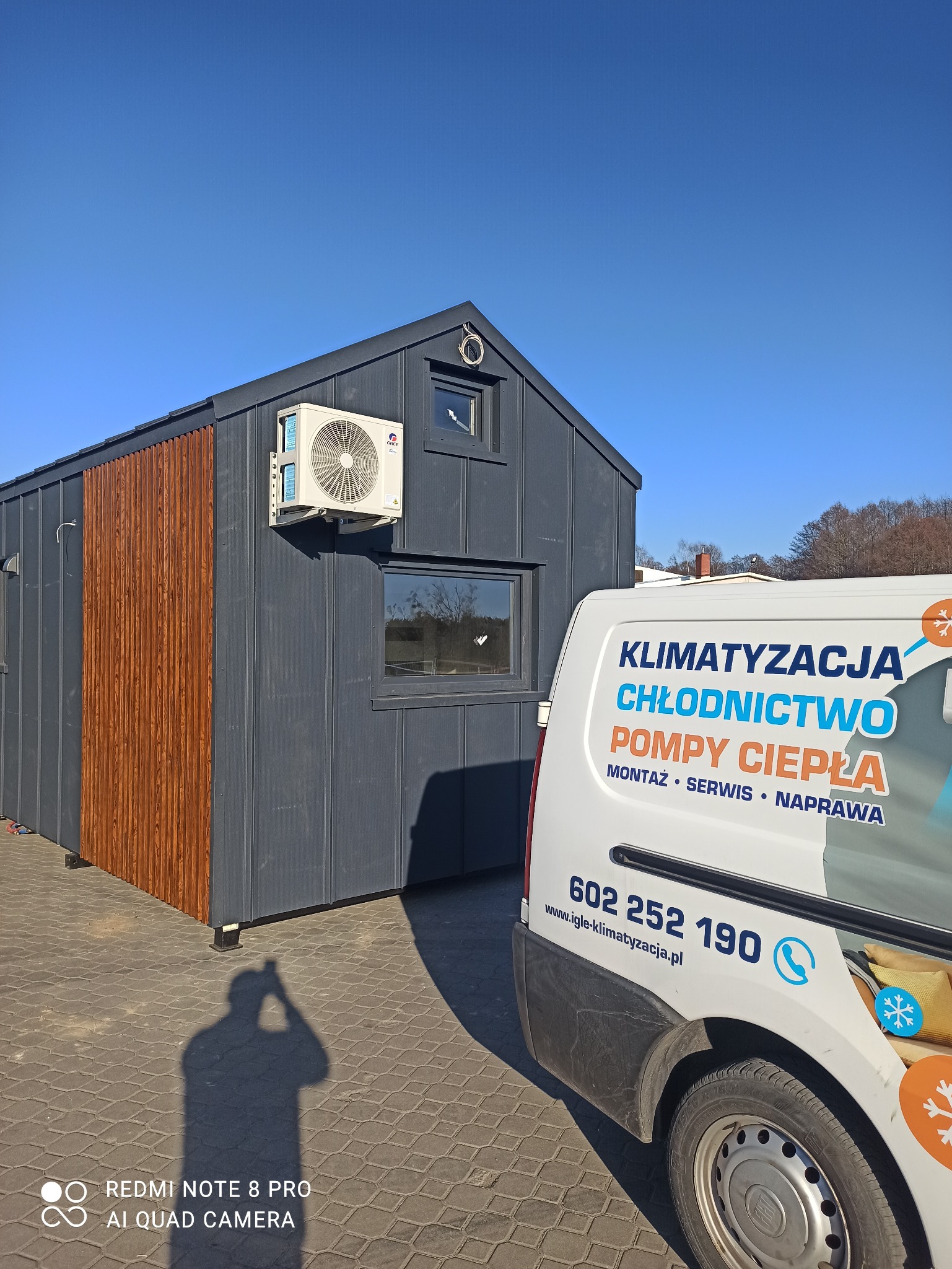 Nowoczesny domek z szarej blachy z drewnianymi akcentami, na którym zamontowano klimatyzator GREE, obok zaparkowany biały samochód serwisowy z reklamą firmy zajmującej się klimatyzacją i pompami...