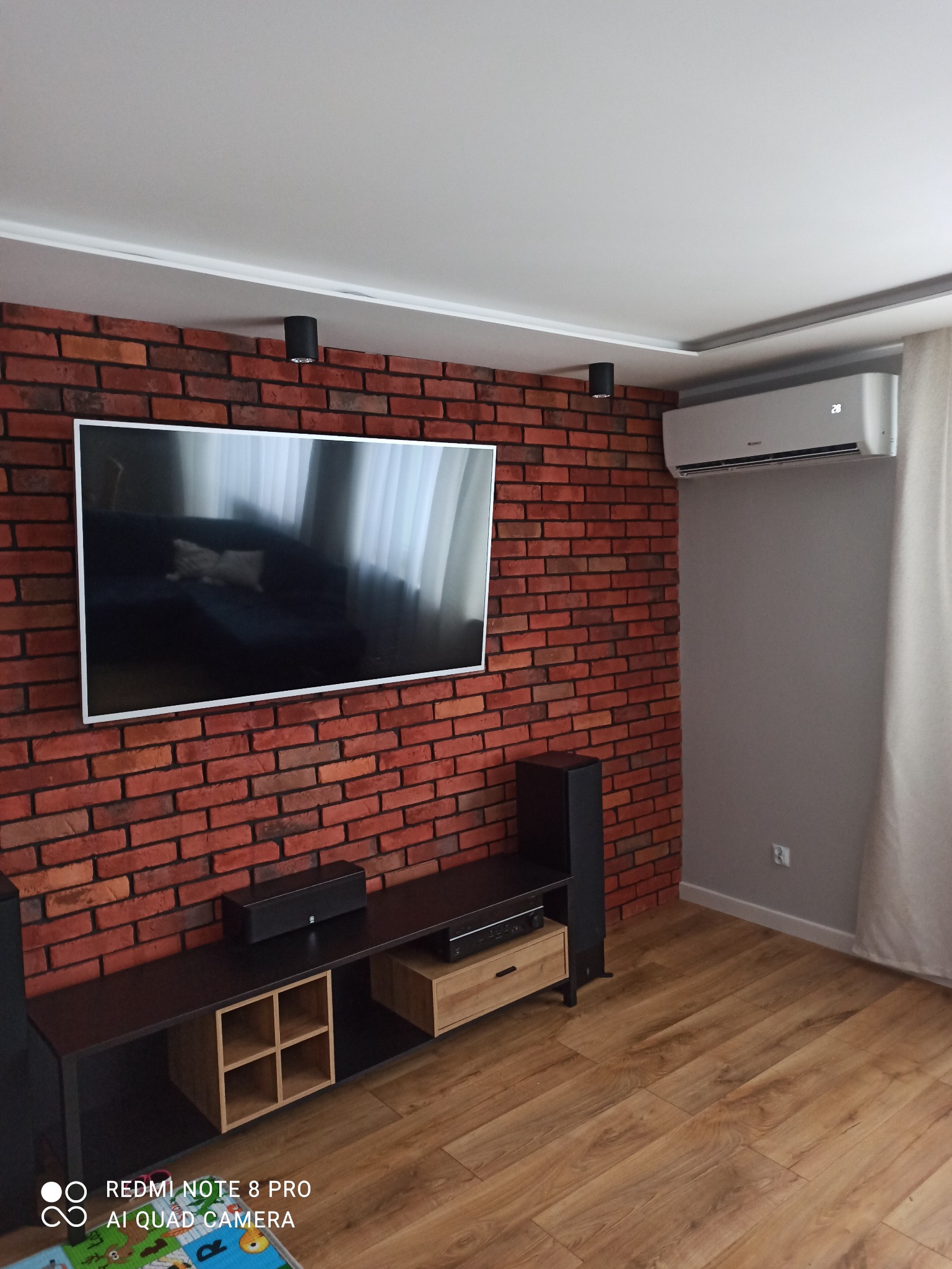 Klimatyzator ścienny zamontowany nad ceglaną ścianą w salonie z drewnianą podłogą, widoczne czarne głośniki i czarna szafka RTV
