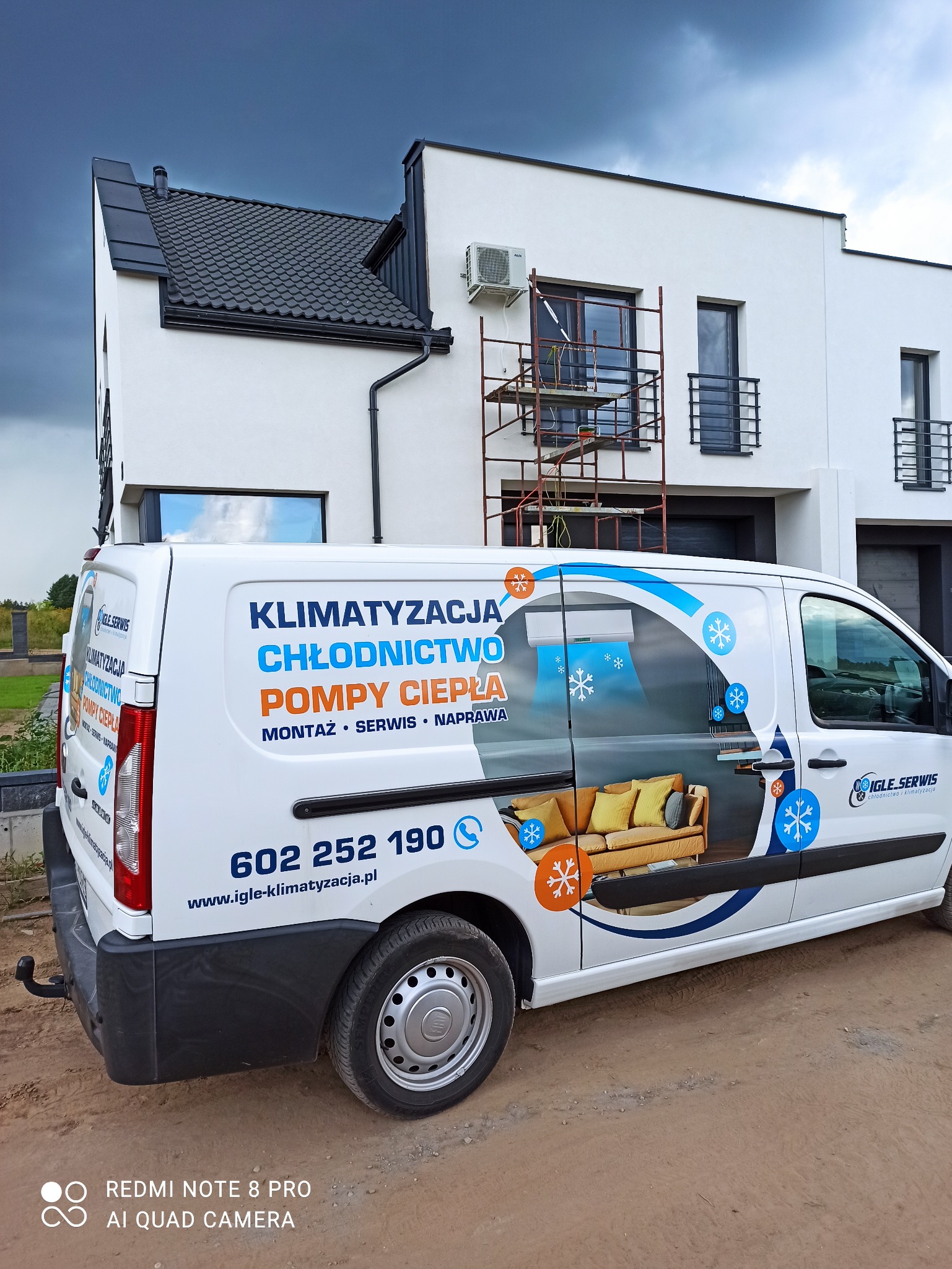 Biały bus serwisowy z reklamą klimatyzacji i pomp ciepła na tle nowoczesnego domu z zamontowaną jednostką zewnętrzną klimatyzacji i rusztowaniem.