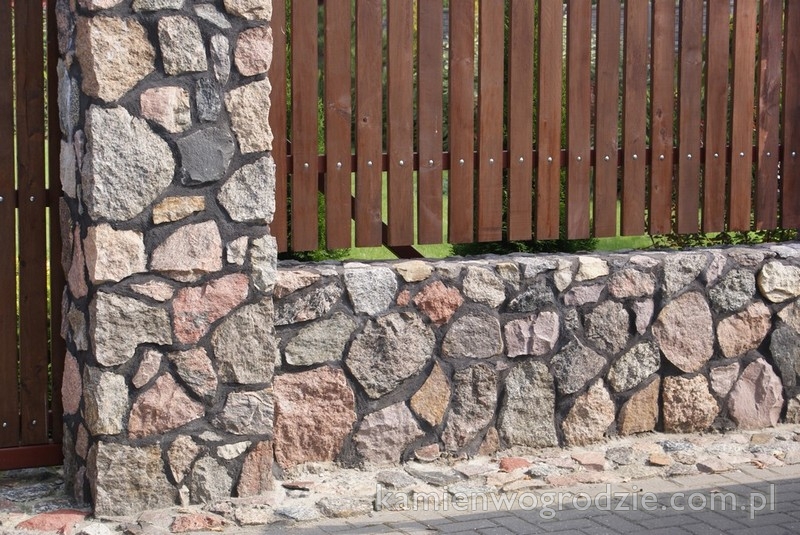 Fragment ogrodzenia z kamienia naturalnego i drewnianych paneli, widoczne nieregularne kształty kamieni w odcieniach szarości i różu, połączonych ciemną zaprawą.