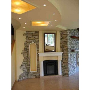 Wnętrze z dekoracyjną ścianą z kamienia, imitacją kominka z białym obramowaniem i lustrem, oraz podwieszanym sufitem z oświetleniem punktowym i dwoma podświetlanymi panelami.