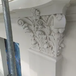 Odrestaurowana, biała, ozdobna kolumna z roślinnymi motywami na elewacji budynku, widoczne rusztowanie w tle.