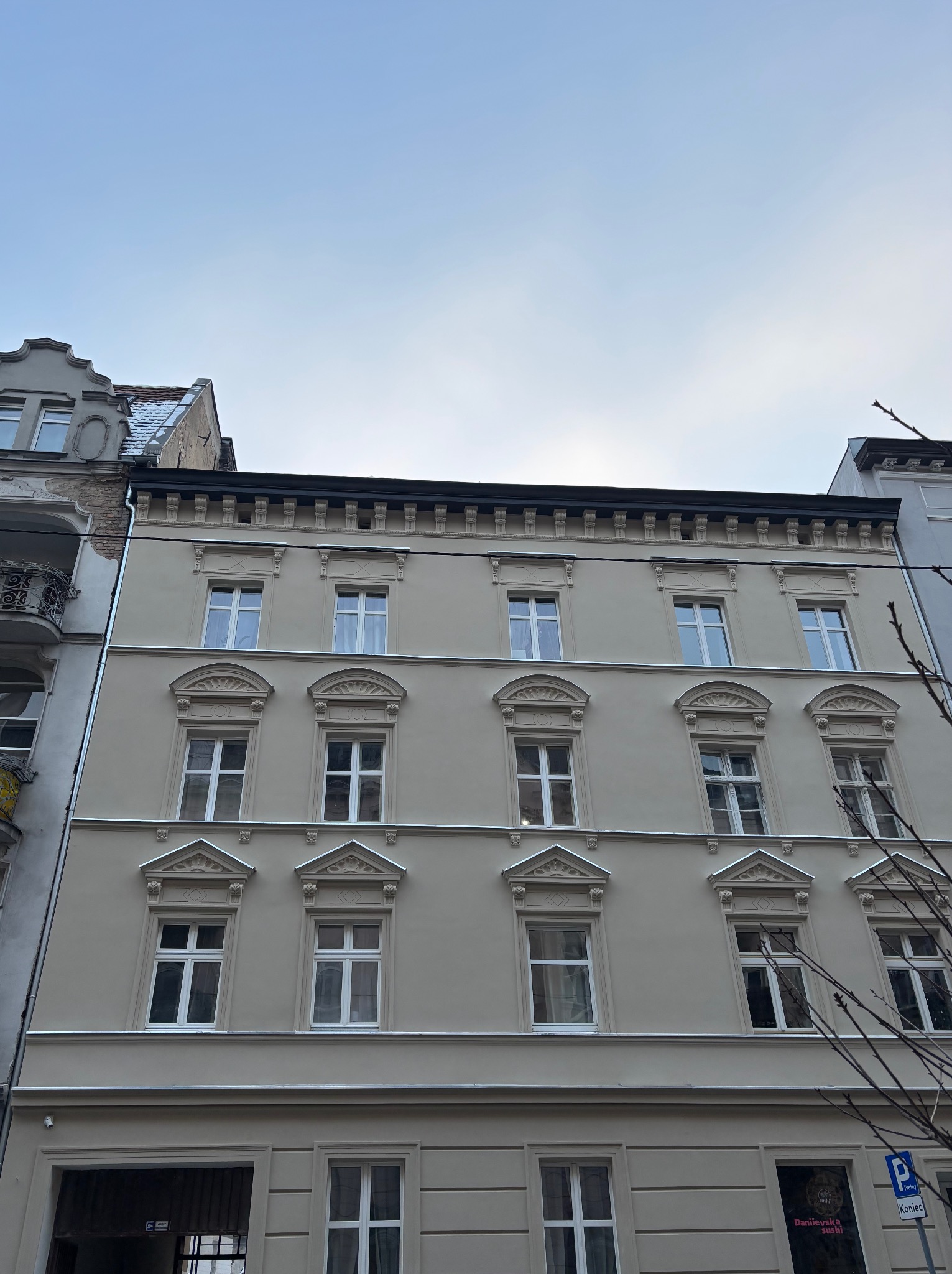 Odnowiona elewacja kamienicy z dekoracyjnymi elementami architektonicznymi wokół okien, pomalowana na jasny kolor. Widok z dołu na budynek.