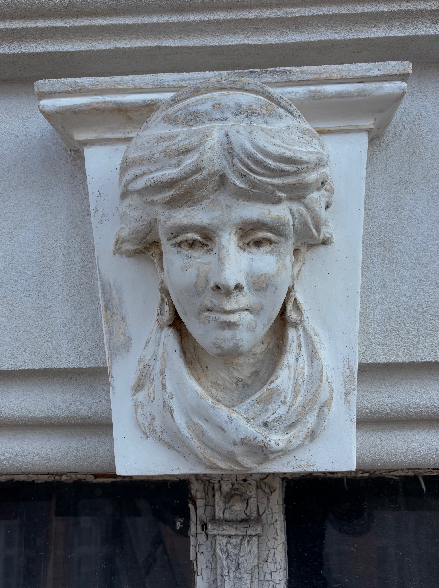 Gipsowa płaskorzeźba kobiecej twarzy z detalami włosów i draperii, umieszczona na elewacji budynku. Widoczne ślady patyny i delikatne uszkodzenia.