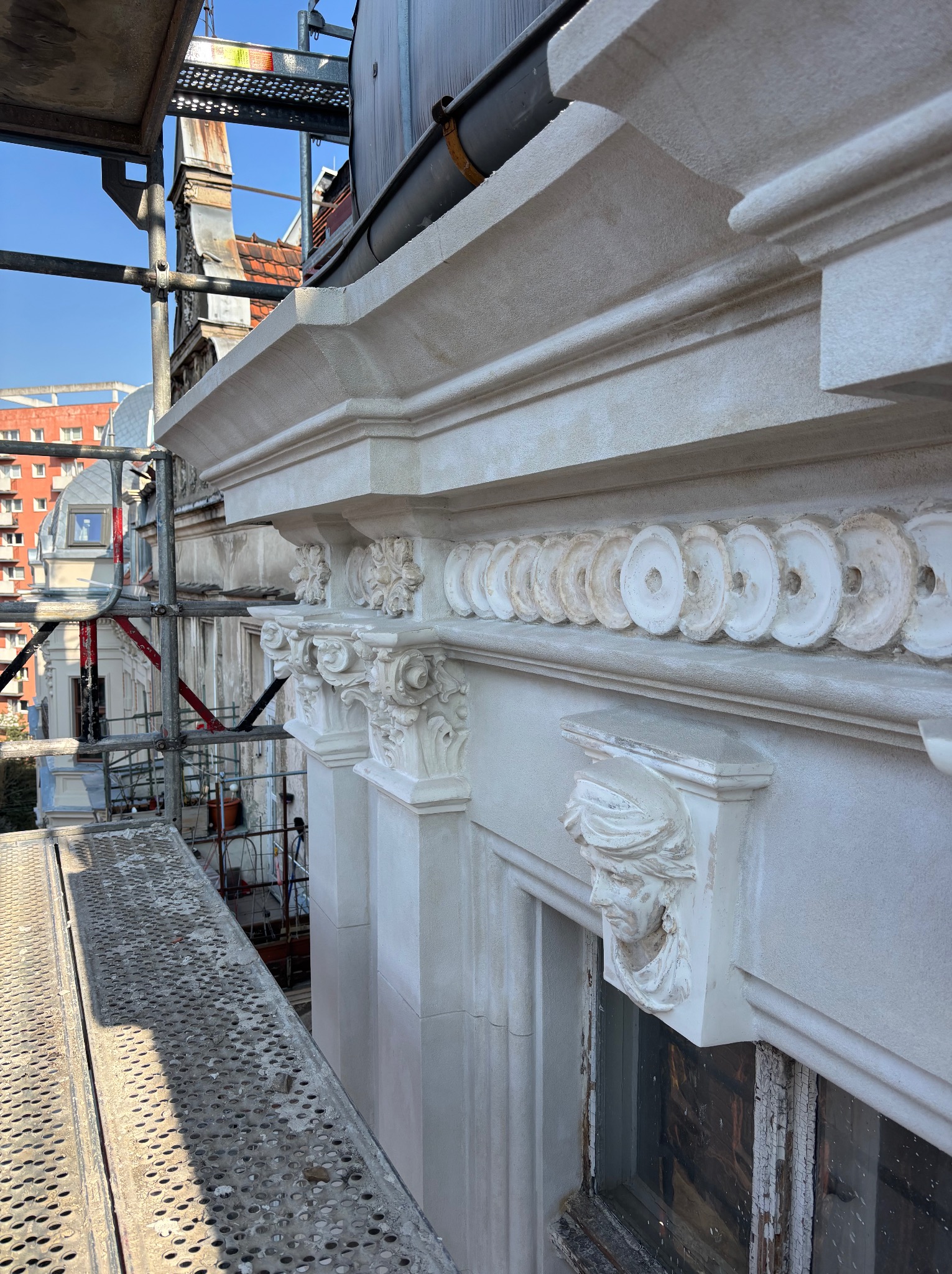 Odrestaurowana fasada kamienicy z detalami sztukaterii, widoczna z rusztowania. Elementy dekoracyjne w postaci płaskorzeźb i ornamentów. Jasna kolorystyka elewacji.