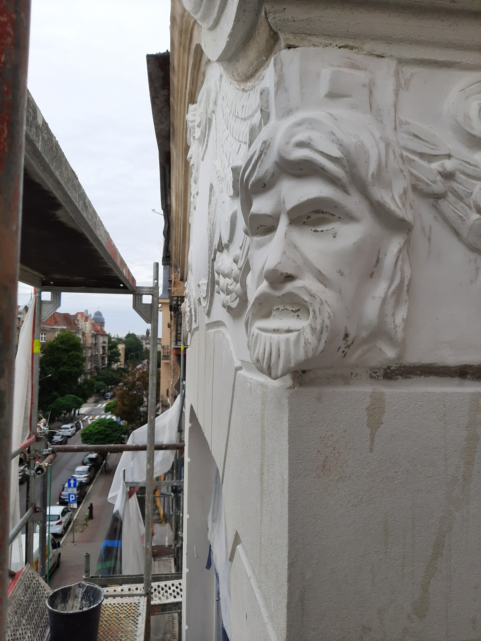 Fragment elewacji kamienicy z rzeźbionym wizerunkiem brodatej męskiej twarzy, widok z rusztowania na ulicę w pochmurny dzień.