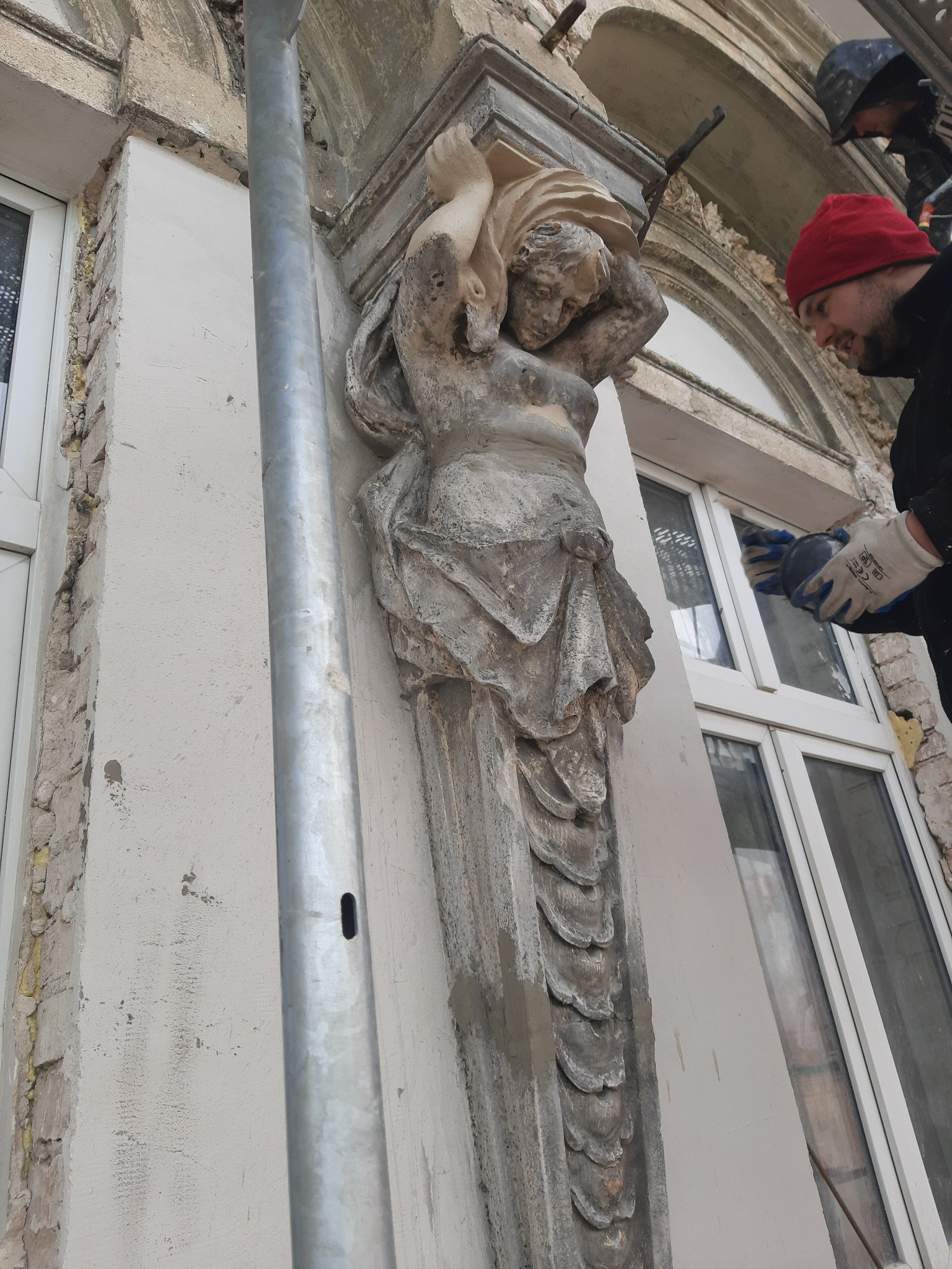 Renowacja elewacji kamienicy: pracownik w czerwonej czapce szlifuje ramę okienną obok kamiennej figury podtrzymującej balkon.
